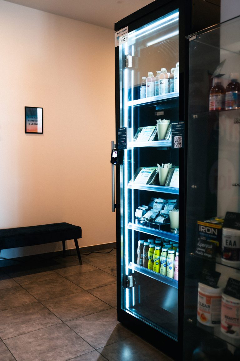 Tall black cabinet with a glass door and lit shelves holding drinks and packaged food