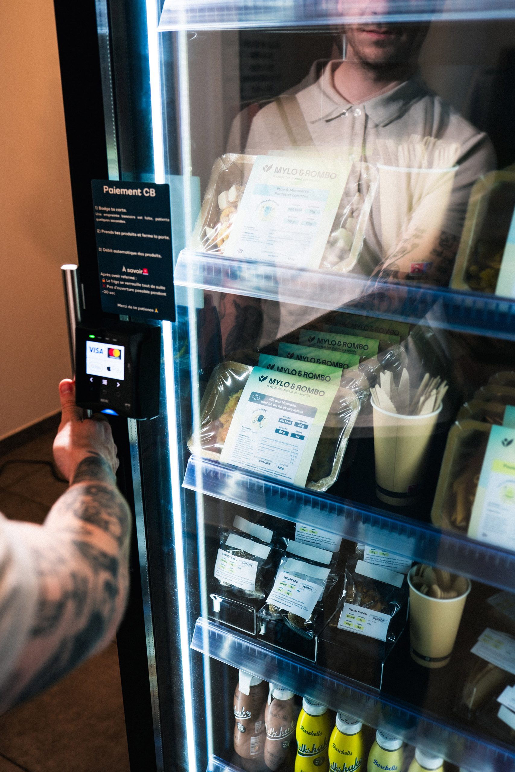 Hand holding a payment terminal at the glass door of a cabinet, shelves of prepared meals and drinks behind the glass
